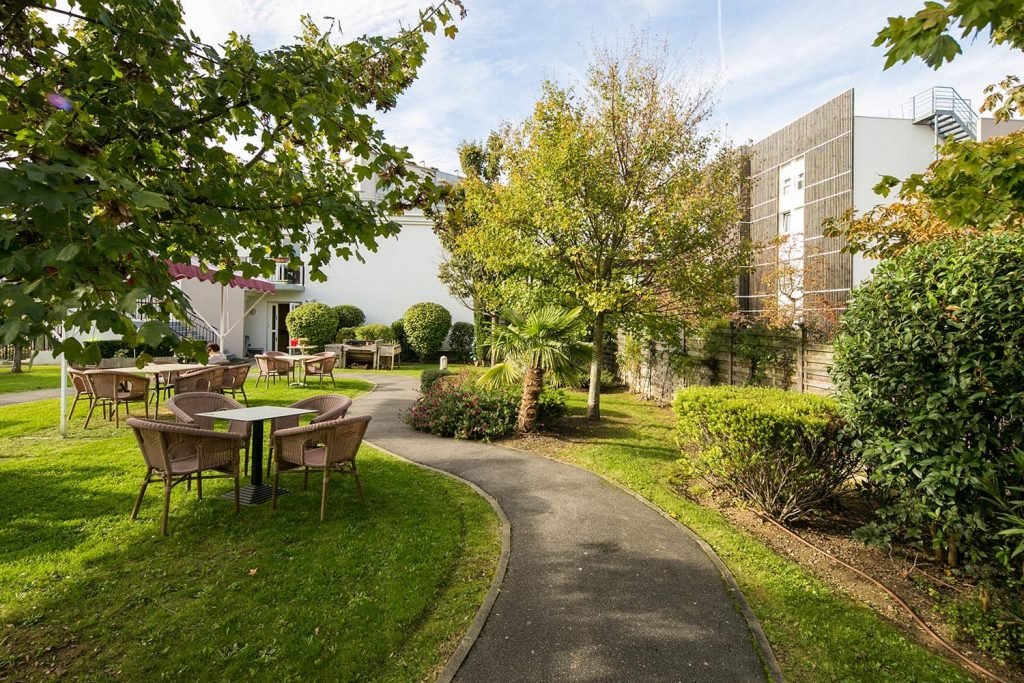 Jardin Sévigné sécurisé avec chemin de marche, tables et chaises sur pelouse et entourées d'arbres